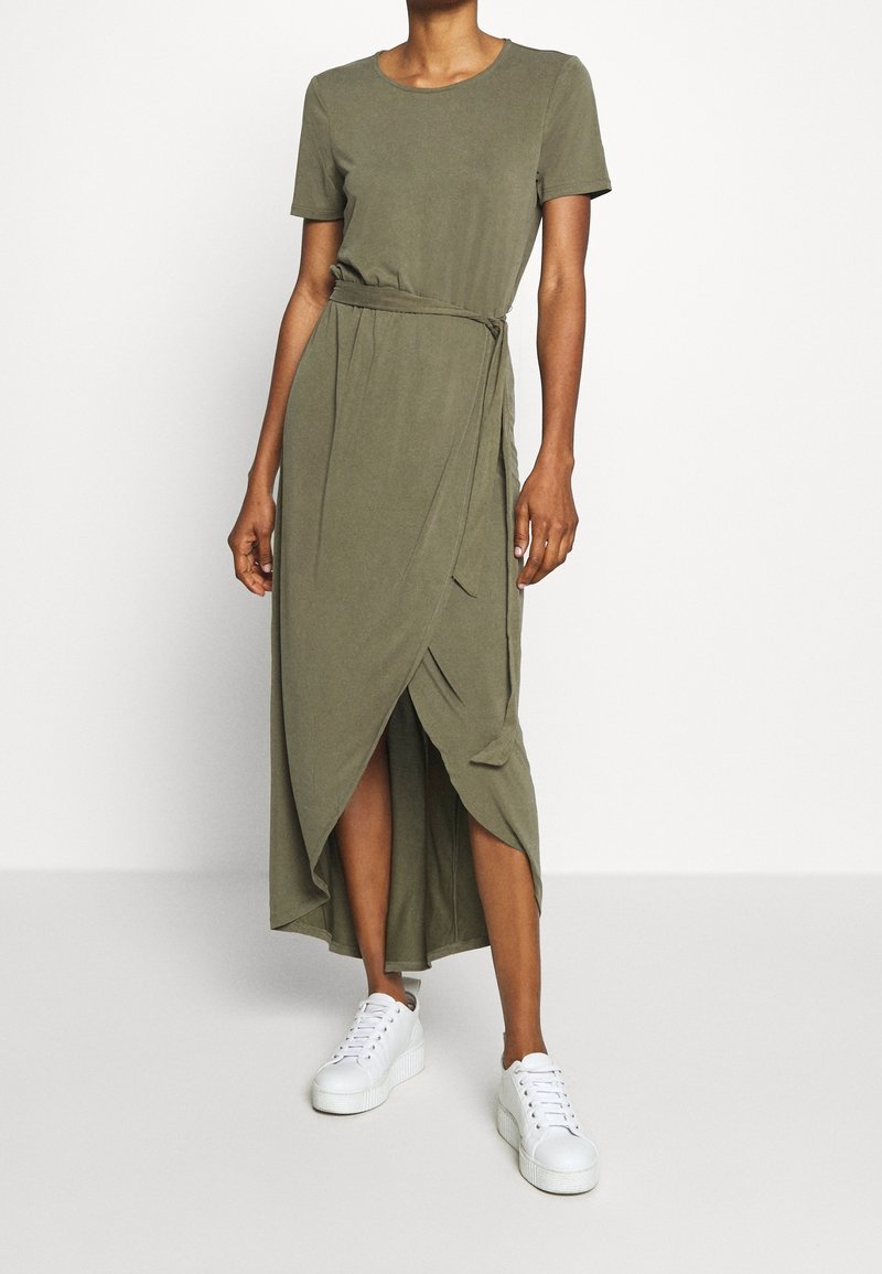 Robe maxi vert olive à manches courtes, taille cintrée et détail cache-cur, avec une fente à l'avant et associée à des baskets blanches.