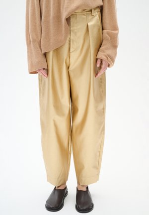 I pantaloni ampi in satin dorato presentano una vita alta, delle pieghe e una texture liscia, abbinati a semplici scarpe nere slip-on.