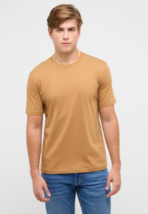 Young man with light brown hair wearing a plain tan short-sleeve t-shirt and blue jeans, standing against a plain light background.