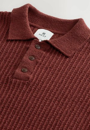 Maranžpunane kootud polo särk, millel on tekstureeritud pind, krae ja kolm tumedat pruunikat nuppu nööbitud alustel. Brändi silt on nähtav.