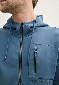 Niebieska bluza zapinana na zamek z kieszenią frontową na zamek, kapturem ze sznurkiem oraz logo "TOM TAILOR" na dole. Wykonana z miękkiego materiału.