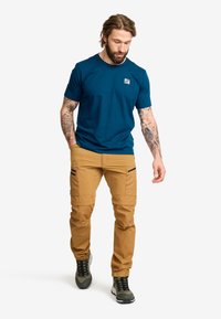 Herrkortärmad skjorta i teal med en logotyp, tillsammans med bruna cargobyxor med dragkedjefickor och mörka kängor med en strukturerad yta.