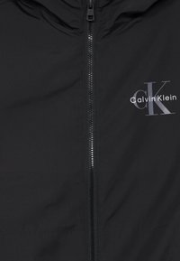 Jachetă neagră cu fermoar din material synthetic neted, cu glugă, fermoar metalic și logo gri pe partea stângă a pieptului.