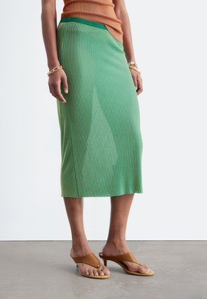 Femme portant une jupe midi côtelée verte, un haut orange, des sandales marron à talons et des bracelets en or et perles, debout sur un sol gris.