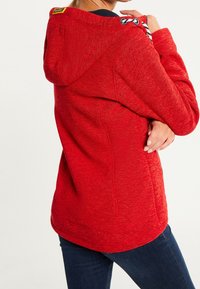 Femme portant une veste à capuche rouge avec des cordons de serrage, vue de dos, se tenant avec un bras croisé sur son torse et la main sur l'épaule.