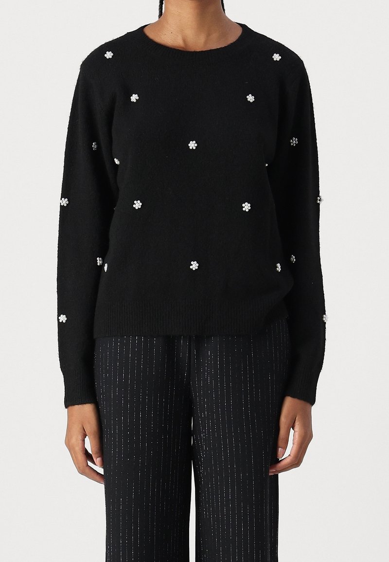 Pull noir à manches longues orné de petits motifs floraux en perles blanches regroupées, porté avec un pantalon noir à fines rayures verticales argentées.