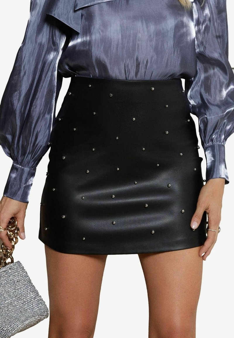 Femme portant une mini-jupe en cuir noire avec de petites clous argentés et un chemisier lavande brillant à manches longues, tenant un sac à main argenté avec une chaîne dorée.