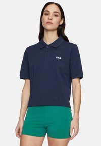 Marineblauwe polo met korte mouwen en kraag, voorzien van vier knopen en een wit logo op de linkerborst. Gekoombineerd met groene shorts.