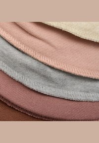 Närbild av staplade mjuka stickade tyger i kräm, rosaskimrande, ljusgrå, mauve och brun med synliga sömmar på de rundade kanterna.