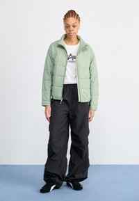 Lys grønn puffy jakke med glidelås foran, kombinert med løse svarte bukser og svarte joggesko. Modellen står mot en ensfarget bakgrunn.