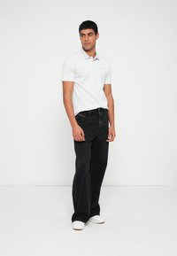 Bijela polo majica s kratkim rukavima, uparena s crnim hlačama širokih nogavica i svijetlim cipelama. Model stoji ispred jednobojne pozadine.