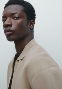 Jeune homme noir avec des cheveux courts et bouclés, portant une veste beige et une chemise sombre, regardant sur le côté contre un fond uni clair.