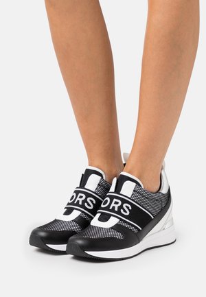 Piedi che indossano sneaker slip-on nere e bianche con tomaia in mesh decorato e ampie fasce elastiche sulla parte superiore con la scritta "ORS".