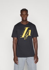 Musta puuvillainen t-paita, jossa on iso keltainen "LA"-logo liikettä kuvaavilla viivoilla sekä pieni Nike-swoosh. Pyöreä pääntie, lyhyet hihat, normaali istuvuus.