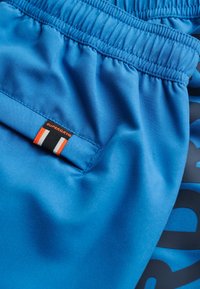 Shorts de sport bleus en tissu lisse avec une taille élastique, dotés d'une petite étiquette noire avec des accents orange et blanc.