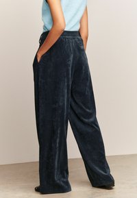 Marineblauwe corduroy wijde broek met een elastische tailleband, zijzakken en een textuurstof, getoond vanuit een achteraanzicht.