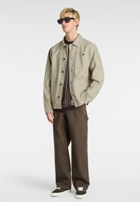 Light beige jacket with a plain design, brown ribbed sweater underneath, wide brown trousers, and black sunglasses. Textured materials are visible.