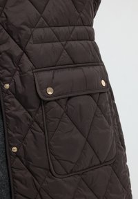 Gequiltete bruine vest met een borstzak met een drukknopsluiting en gouden hardware, met een gladde, lichtgewicht stofstructuur.