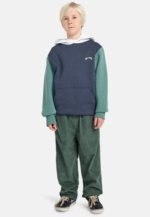 Marineblauer und türkisfarbener Farbblock-Hoodie mit einer Fronttasche, ausgestattet mit einem weißen Logo. Kombiniert mit grünen Cordhosen und schwarzen Sneakers.
