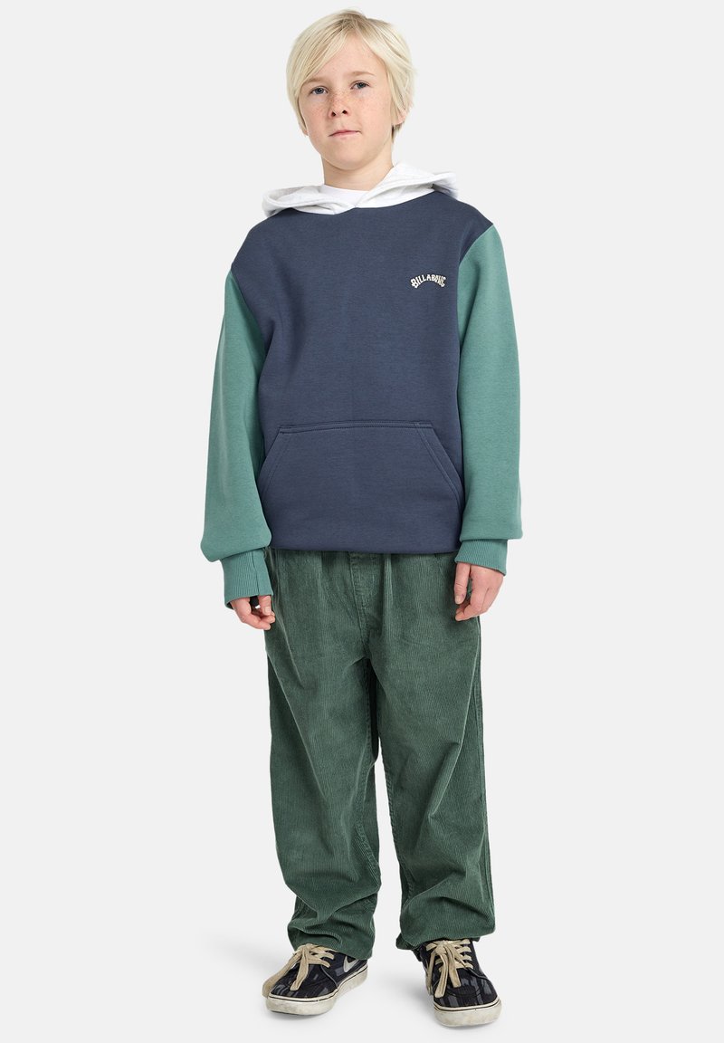 Marineblauwe en teal kleurgebroken hoodie met een voorzak, voorzien van een wit logo. Gecombineerd met groene corduroy broek en zwarte sneakers.