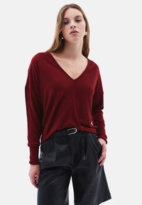 Maglione bordeaux a maglia con scollatura a V, vestibilità ampia e maniche lunghe, abbinato a pantaloni corti neri plissettati e una cintura con fibbia in argento.