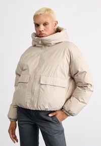 Veste coupe-vent beige avec capuche, fermeture éclair �à l'avant et deux poches avant. Présente une texture légèrement brillante et lisse ainsi que des poignets élastiques.