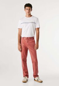 T-shirt bianca a maniche corte con righe blu e logo abbinata a pantaloni di jeans rossi e sneaker beige con accenti neri.