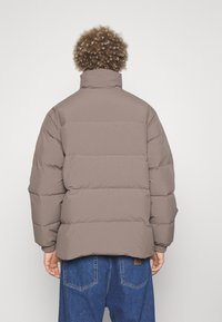 Brown puffer jacket with horizontal seams, high collar, and ribbed cuffs. Worn with blue denim jeans. Simple, functional design.
