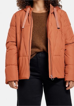 Vrouw in een roestkleurige gewatteerde jas met een rits over een bruine trui en zwarte hoogblijvende broek, staand met ontspannen handen.