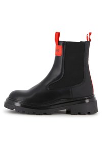 Botim de tornozelo em couro preto com sola robusta, apresentando painéis laterais elásticos vermelhos e uma aba para facilitar o calçar. Textura lisa e design com biqueira arredondada.