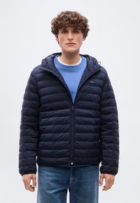 Νέος άνδρας με σγουρά μαλλιά που φοράει ναυτικό μπουφάν με κουκούλα και καπιτονέ επένδυση πάνω από μπλε φούτερ και μπλε τζιν, με φόντο ένα απλό ανοιχτόχρωμο τοπίο.