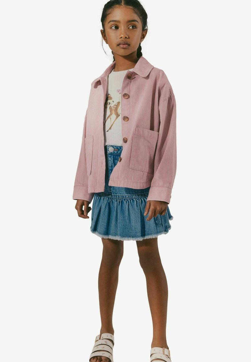 Veste en velours côtelé rose avec des boutons en bois, t-shirt graphique blanc, jupe en denim superposée et sandales beiges ; combinaison de tenue décontractée et ludique.