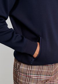 Mornarska dukserica s rebrastim rukavima i bočnom zip džepom, u kombinaciji s smešnim kariranim hlačama u smešnim tonovima crvene, žute i bele boje.