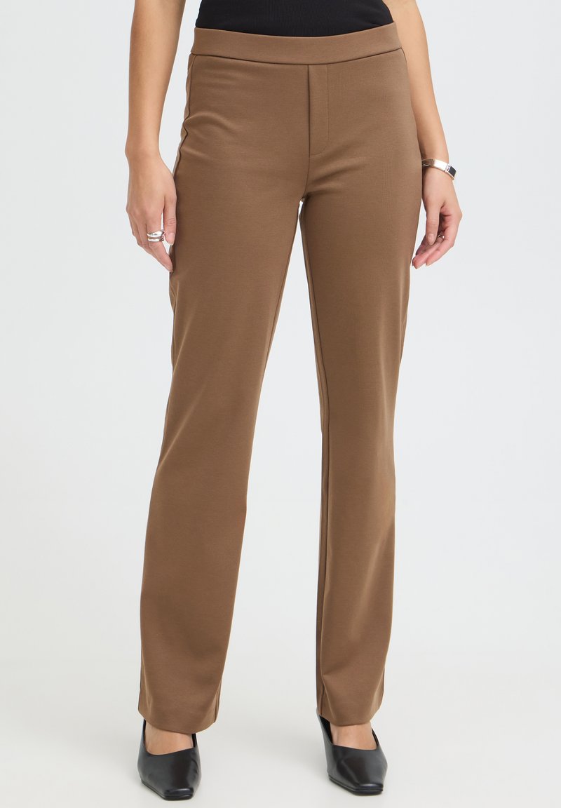 Femme portant un pantalon marron droit taille haute, associé à des chaussures noires à bouts pointus et un haut noir.