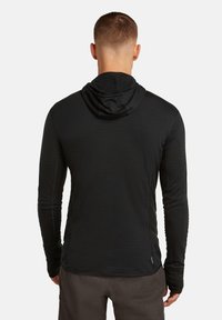 Hombre visto desde atrás, vistiendo una sudadera negra de manga larga y pantalones gris oscuro, contra un fondo claro y sencillo.