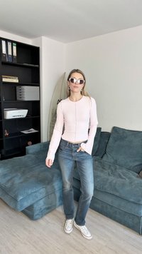 Világos rózsaszín, hosszú ujjú bordázott felső, kerek nyakkal, a kék farmernadrággal és fehér tornacipővel párosítva. Háttérben egy kék kanapé látható.