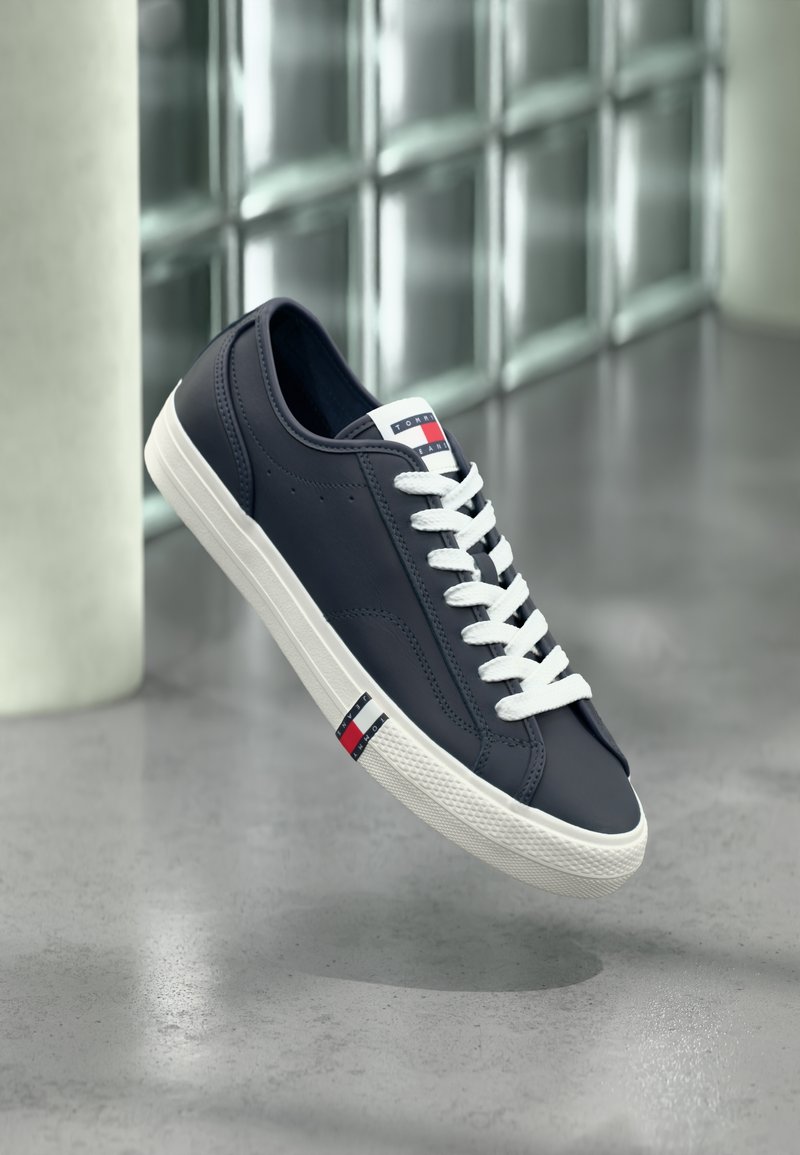 Tumesinine tennisking valgete paelte ja tallaga, millel on Tommy Hilfigeri logo keelel ja küljel, hõljumas siseruumide halli põranda kohal.