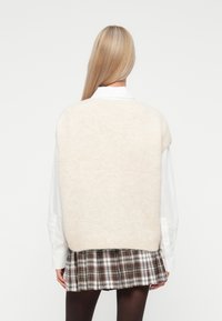 Blondynka tyłem, ubrana w biały, oversize'owy sweter bez rękawów na białej koszuli oraz w brązową, w kratę spódnicę plisowaną i czarne rajstopy.