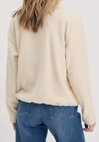 Jersey de forro polar de color crema con un ajuste holgado, mangas largas y una textura sutil. Combinado con vaqueros azules, mostrando detalles traseros.