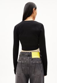 Schwarzes Langarmshirt kombiniert mit grauen Jeans, die ein gelbes Etikett am Bund und zwei Gesäßtaschen haben. Glattes Material mit taillierter Passform.