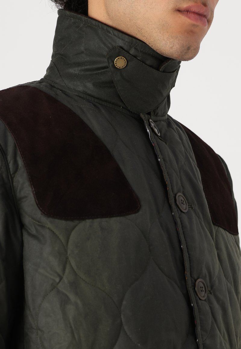 Grüne Steppjacke mit hohem Kragen, braunen Wildleder-Akzenten an den Schultern und Druckknopfverschlüssen. Sie verfügt über eine strukturierte, matte Oberfläche.