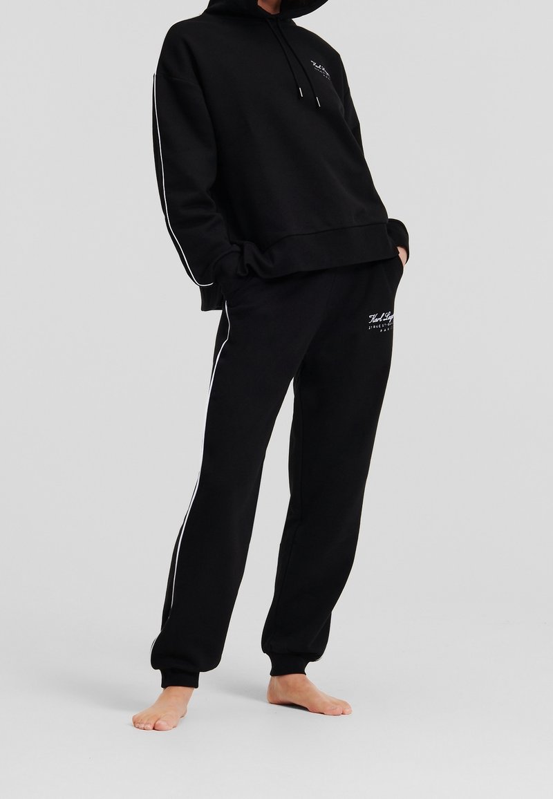 Personne portant un sweat à capuche noir et un pantalon de survêtement noir avec des bandes blanches sur les côtés et un logo, debout pieds nus sur un fond uni.