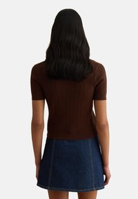 Mujer con cabello largo y negro que lleva un suéter marrón de manga corta con textura y una falda vaquera azul, vista desde atrás contra un fondo liso.