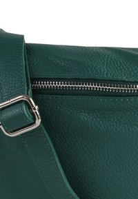 Turquoise leren handtas met een gestructureerd oppervlak, voorzien van een zilveren rits en een metallic gespband, die het strakke ontwerp benadrukt.