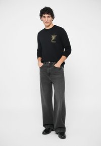 Schwarzes Langarmshirt mit goldener Grafik, kombiniert mit lockeren grauen Denim-Jeans. Das Model hat lockige Haare, die Hände in den Taschen, vor einem schlichten Hintergrund.