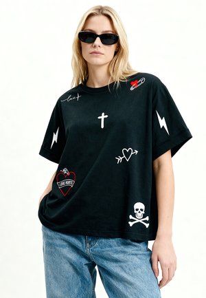 Jeune femme aux cheveux blonds portant des lunettes de soleil noires, un t-shirt noir oversize avec des imprimés graphiques blancs et rouges, et un jean bleu.