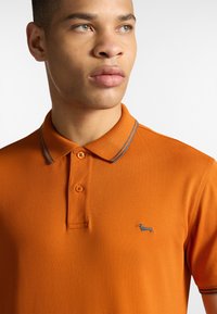 Orange polo t-shirt med tekstureret finish, der har en to-knaps placket, grå stribede kraveaccenter og et lille broderet logo.
