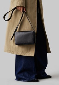 Black leather crossbody bag with a rectangular shape, embossed logo, and adjustable strap, held by a person in a beige coat and dark jeans.