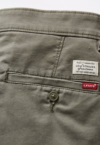 Bawełniana tkanina oliwkowozielona z tylną kieszenią, wyposażona w metkę Levi's i guzik z kwiatowym wzorem. Widoczne są podwójne szwy.