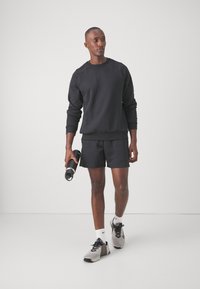 Sweatshirt noir à col rond, shorts assortis et chaussures de sport grises. Le mannequin tient une bouteille d'eau noire, présentant une texture lisse.